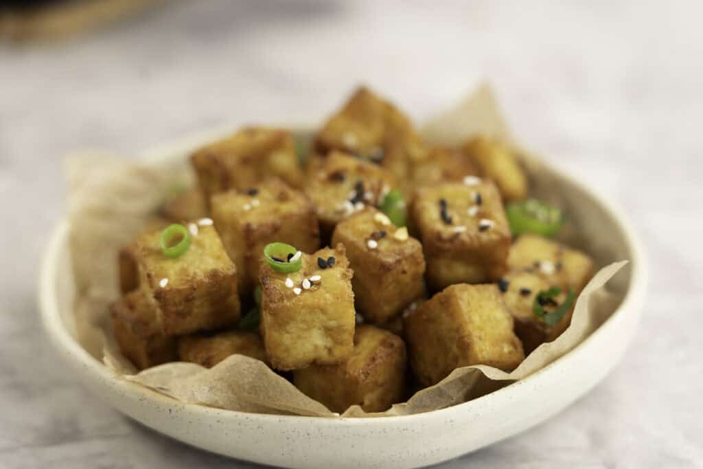 A small plate with crispy cubes of golden brown tofu garnished with sesame seeds and spring onion.