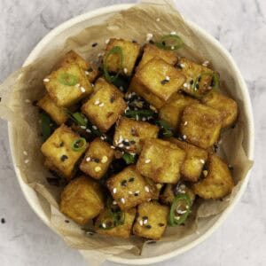 A small plate with crispy cubes of golden brown tofu garnished with sesame seeds and spring onion.