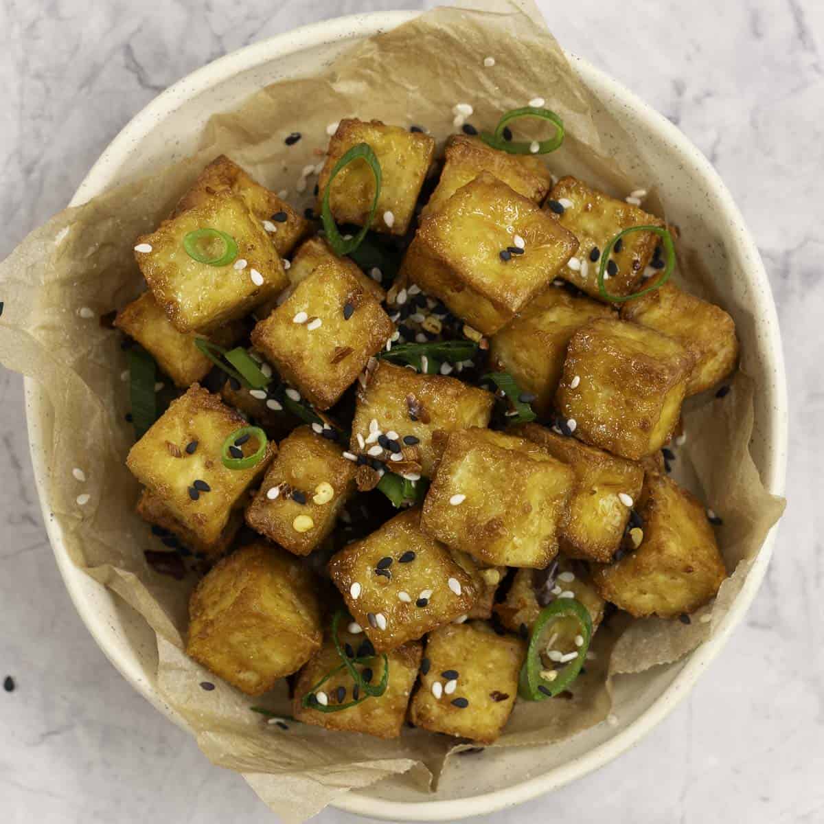A small plate with crispy cubes of golden brown tofu garnished with sesame seeds and spring onion.