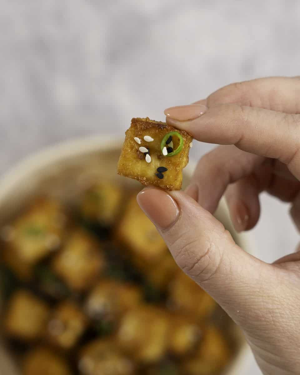 Womans hand holding a crispy golden brown cube of tofu with sesame seeds and spring onion.