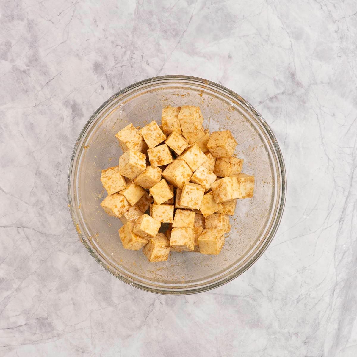 Tofu pieces cut into small cubes in a large glass bowl on marble benchtop with seasoning sauce mixture inside.