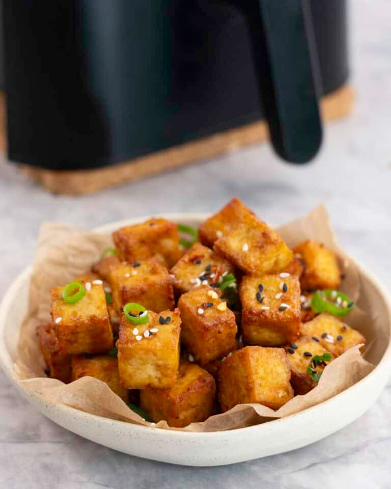 A small plate with crispy cubes of golden brown tofu garnished with sesame seeds and spring onion.