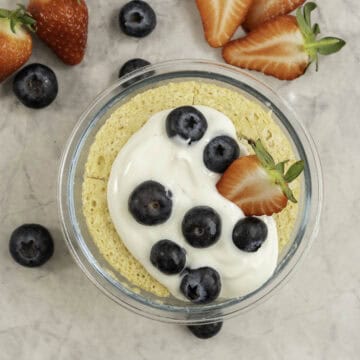 Cooked pancake in a small glass ramekin topped with yogurt, blueberries and strawberries. Fruit scattered on benchtop.