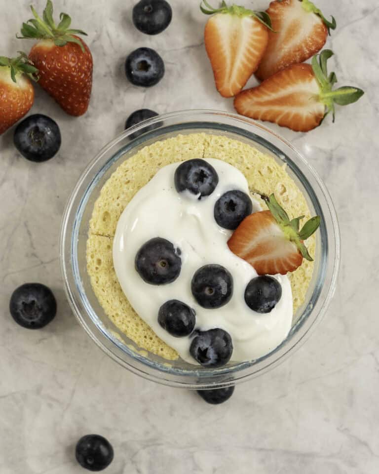 Cooked pancake in a small glass ramekin topped with yogurt, blueberries and strawberries. Fruit scattered on benchtop.