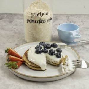 Two stacked pancakes with topped with yogurt, blueberries and strawberries, tall glass jar labelled "protein pancake mix" on benchtop.