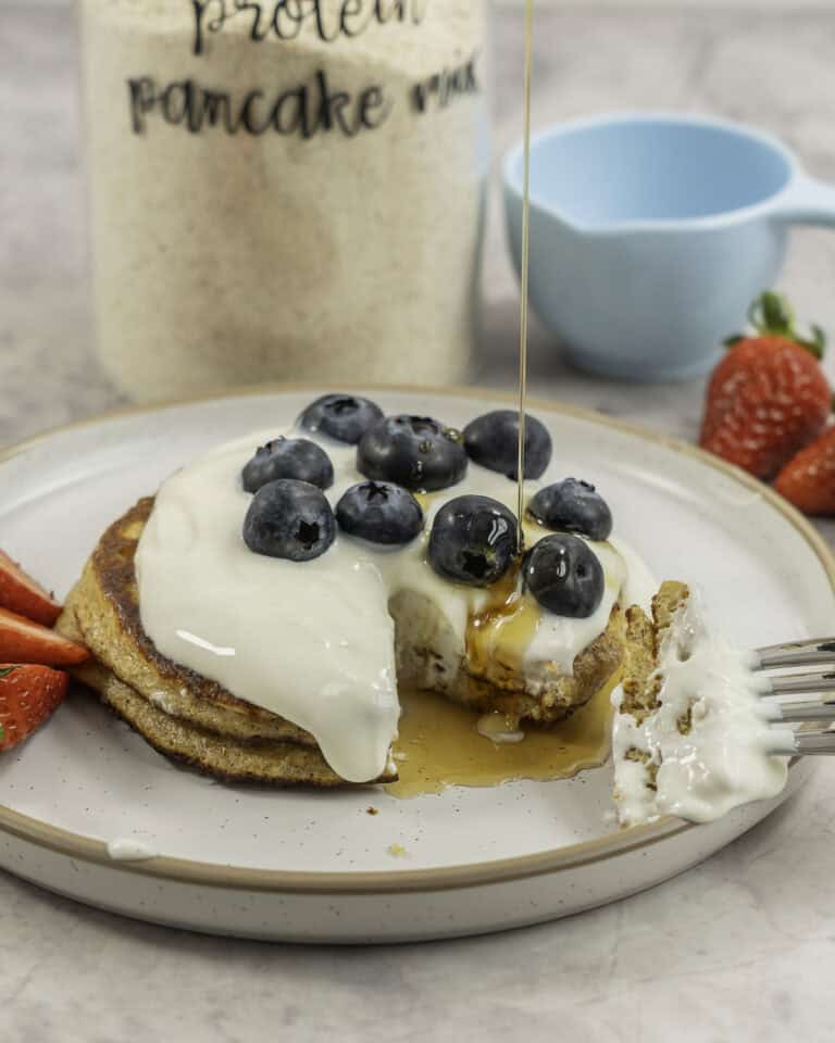 Two stacked pancakes with topped with yogurt, blueberries and strawberries, maple syrup drizzling on top, tall glass jar labelled "protein pancake mix" on benchtop.