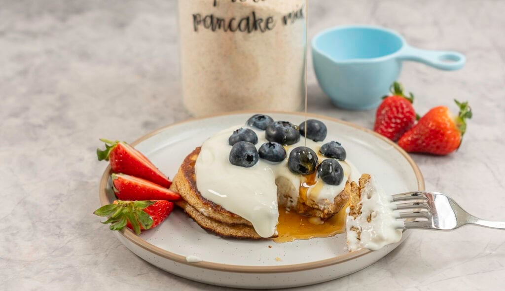 Two stacked pancakes with topped with yogurt, blueberries and strawberries, maple syrup drizzling on top, tall glass jar labelled "protein pancake mix" on benchtop.
