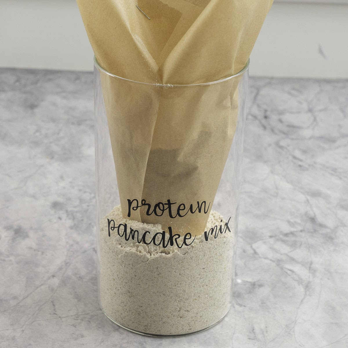Tall glass container labelled "protein pancake mix" with brown baking paper inside used as a funnel, quarter filled with pancake mix.