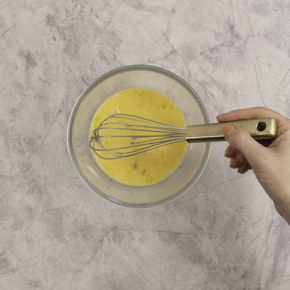 Womans hand whisking eggs in a glass bowl on benchtop.
