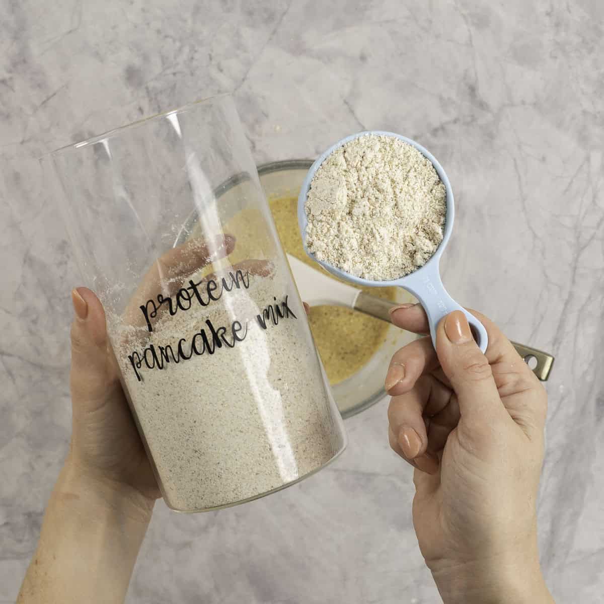Woman's hands holding a tall glass jar labelled "protein pancake mix" and a measuring cup with mix inside.