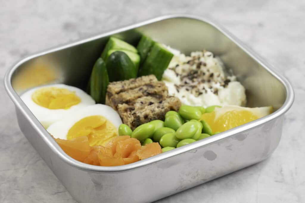 Stainless steel container with chopped cucumber, bread pieces, salmon, cottage cheese a hard boiled egg sliced in half, edamame beans and cottage cheese with a slice of lemon inside.