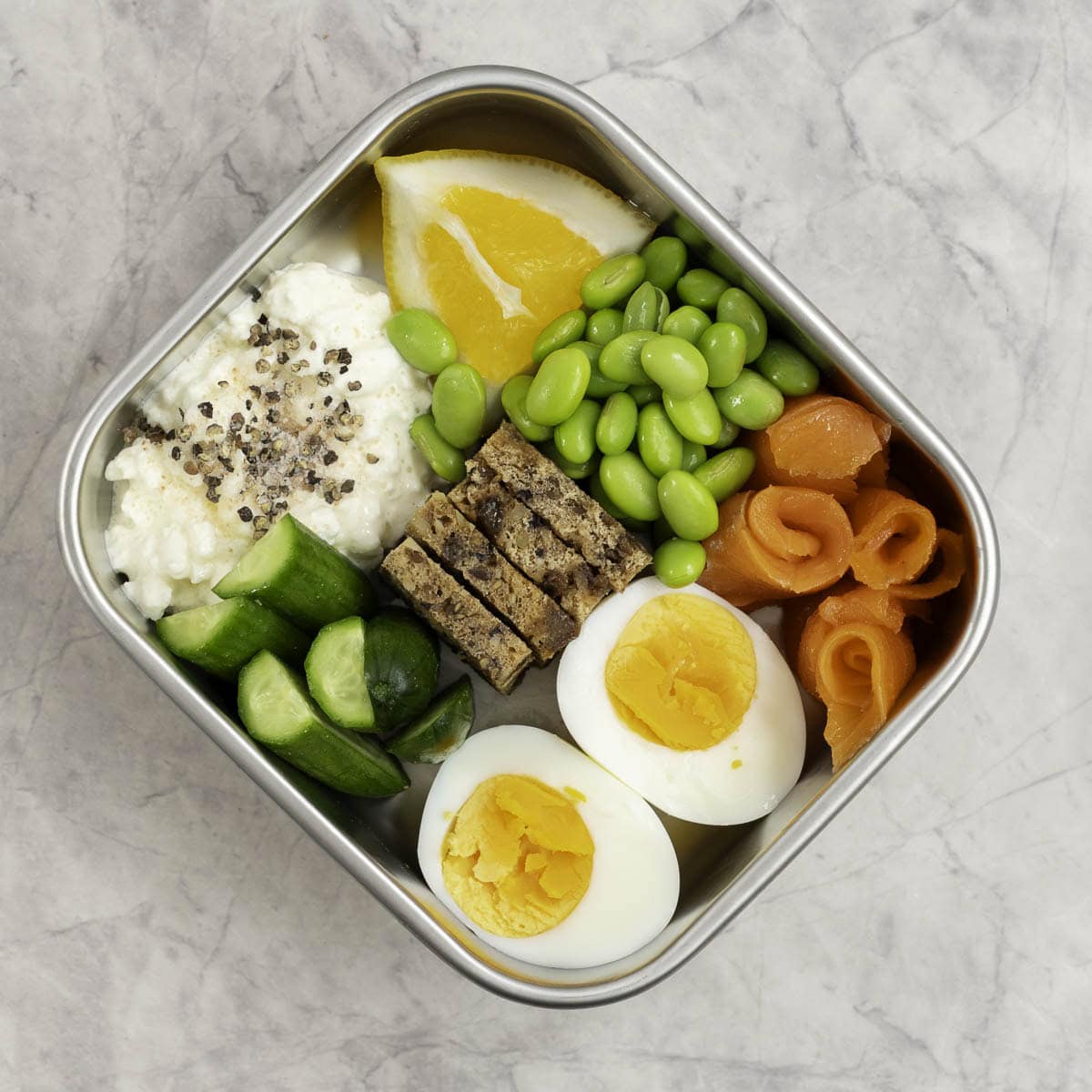 Stainless steel container with chopped cucumber, bread pieces, cottage cheese a hard boiled egg sliced in half, edamame beans and cottage cheese with a slice of lemon inside.
