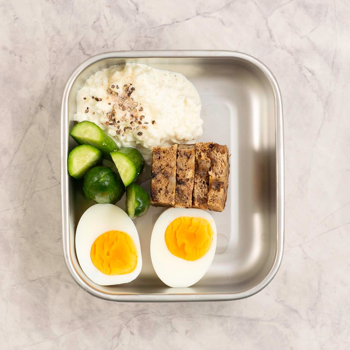 Bento box, half packed with cottage cheese, cucumber, boiled egg, salt and pepper visible.