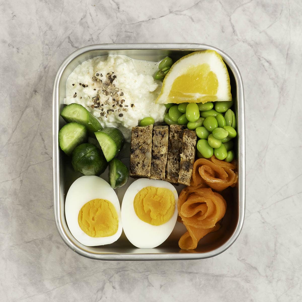 Stainless steel container with chopped cucumber, bread pieces, cottage cheese a hard boiled egg sliced in half, edamame beans and cottage cheese with a slice of lemon inside.
