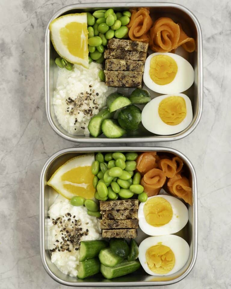 Two stainless steel containers with chopped cucumber, bread pieces, cottage cheese a hard boiled egg sliced in half, edamame beans and cottage cheese with a slice of lemon inside.