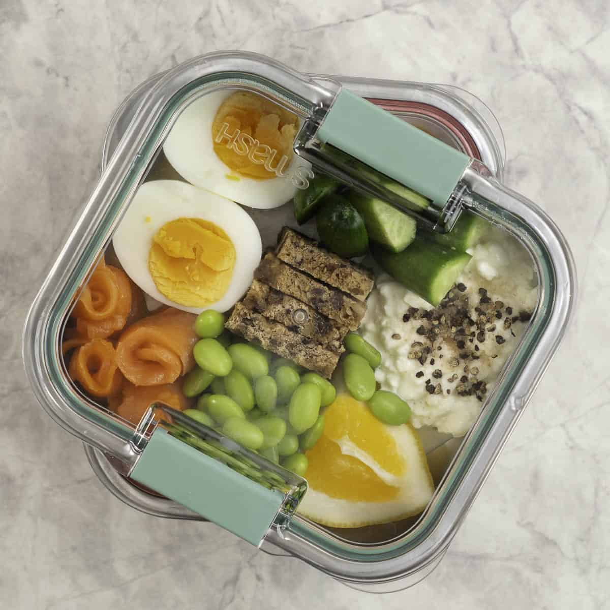 Stainless steel container with chopped cucumber, bread pieces, cottage cheese a hard boiled egg sliced in half, edamame beans and cottage cheese with a slice of lemon inside.
