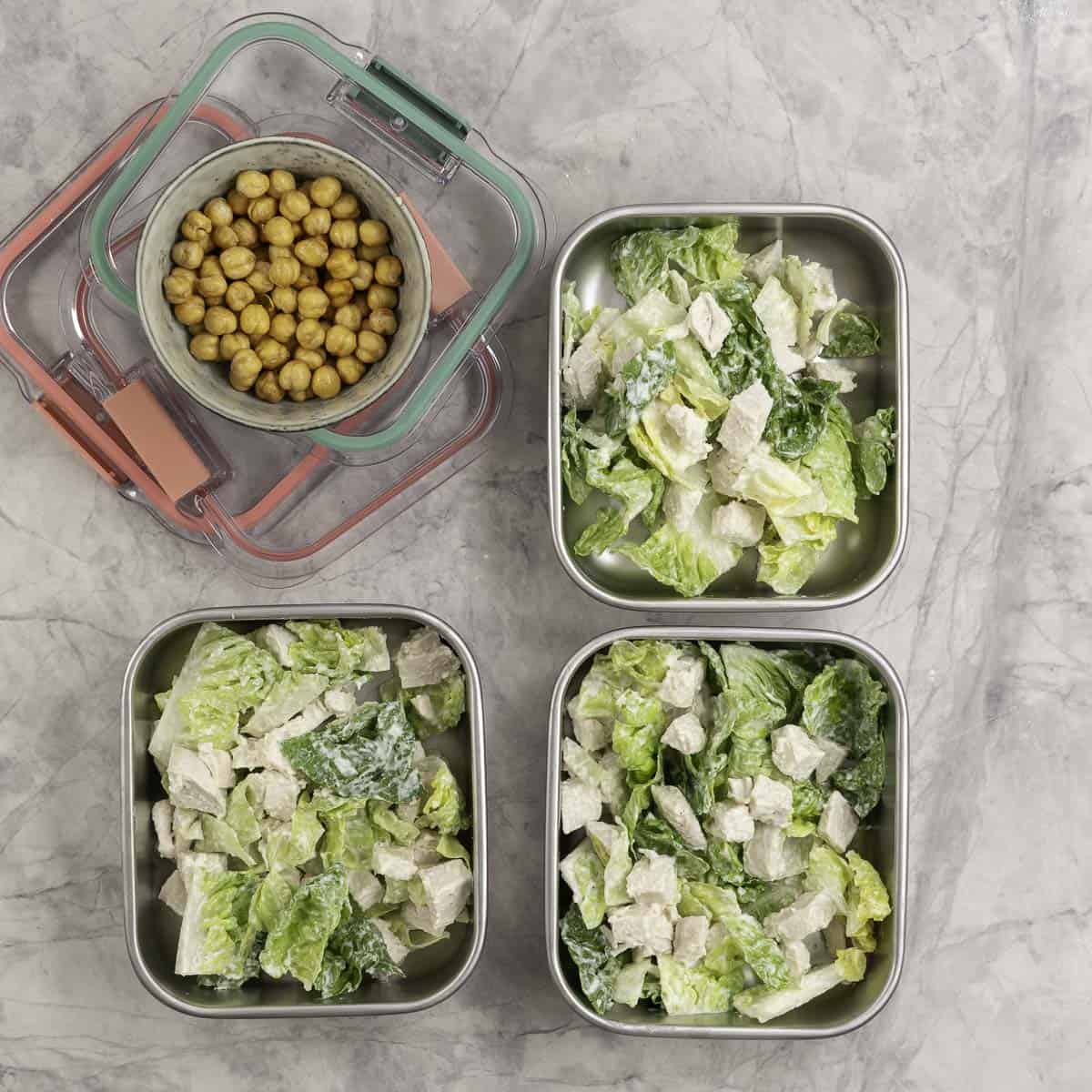 Three containers of chicken caesar salad ready to have their lid put on, next to a separate container of roasted chickpeas. 