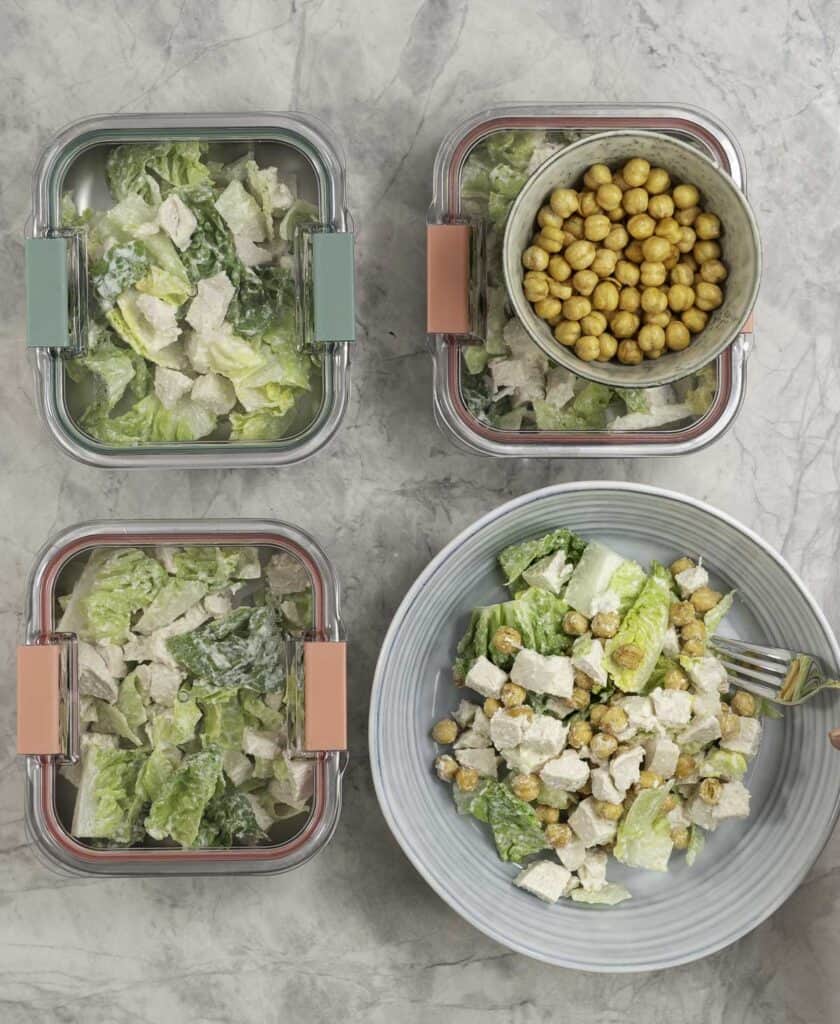 Three containers of chicken caesar salad ready to have their lid put on, plus a blue bowl of salad ready to serve, next to a separate container of roasted chickpeas.