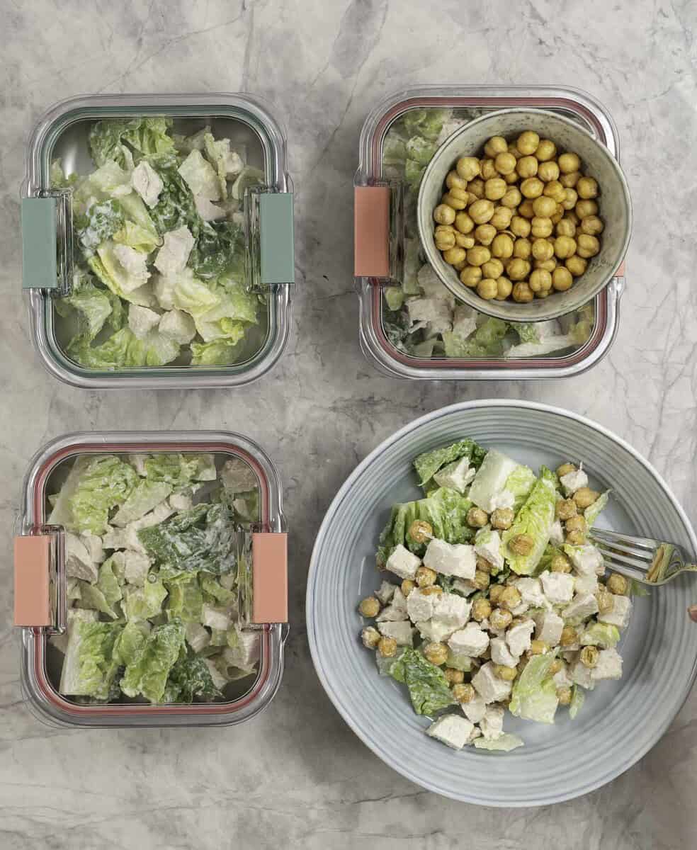 Three containers of chicken caesar salad ready to have their lid put on, plus a blue bowl of salad ready to serve, next to a separate container of roasted chickpeas.