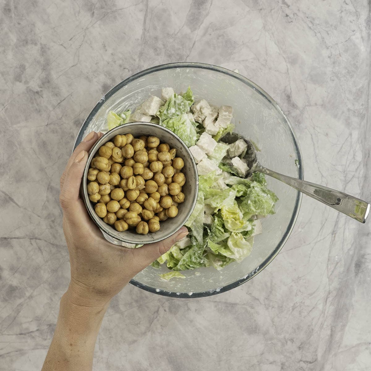A glass bowl of chicken caesar salad with a hand holding a bowl of roasted chicken peas above it. 