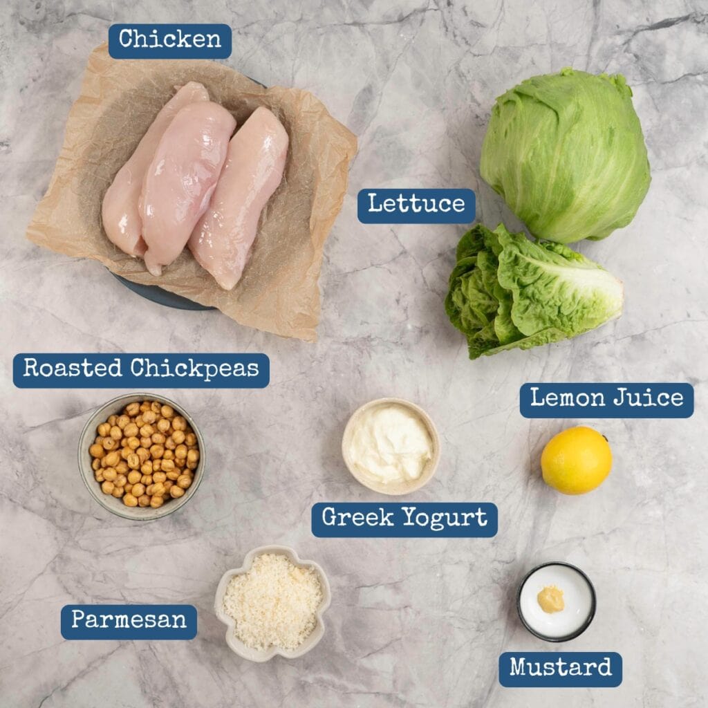 The ingredients to make chicken Caesar Salad laid out on a bench top with text overlay.