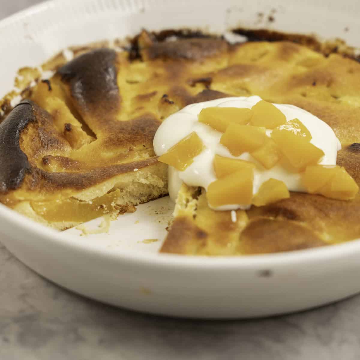 A peach clafoutis with one slice removed, topped with yogurt and more peaches.