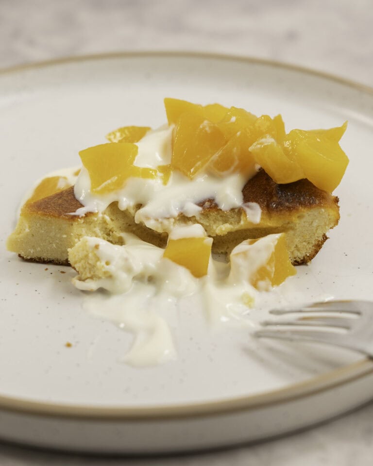 A slice of peach clafoutis on a dessert plate topped with yogurt and diced peaches, with a forkful removed.