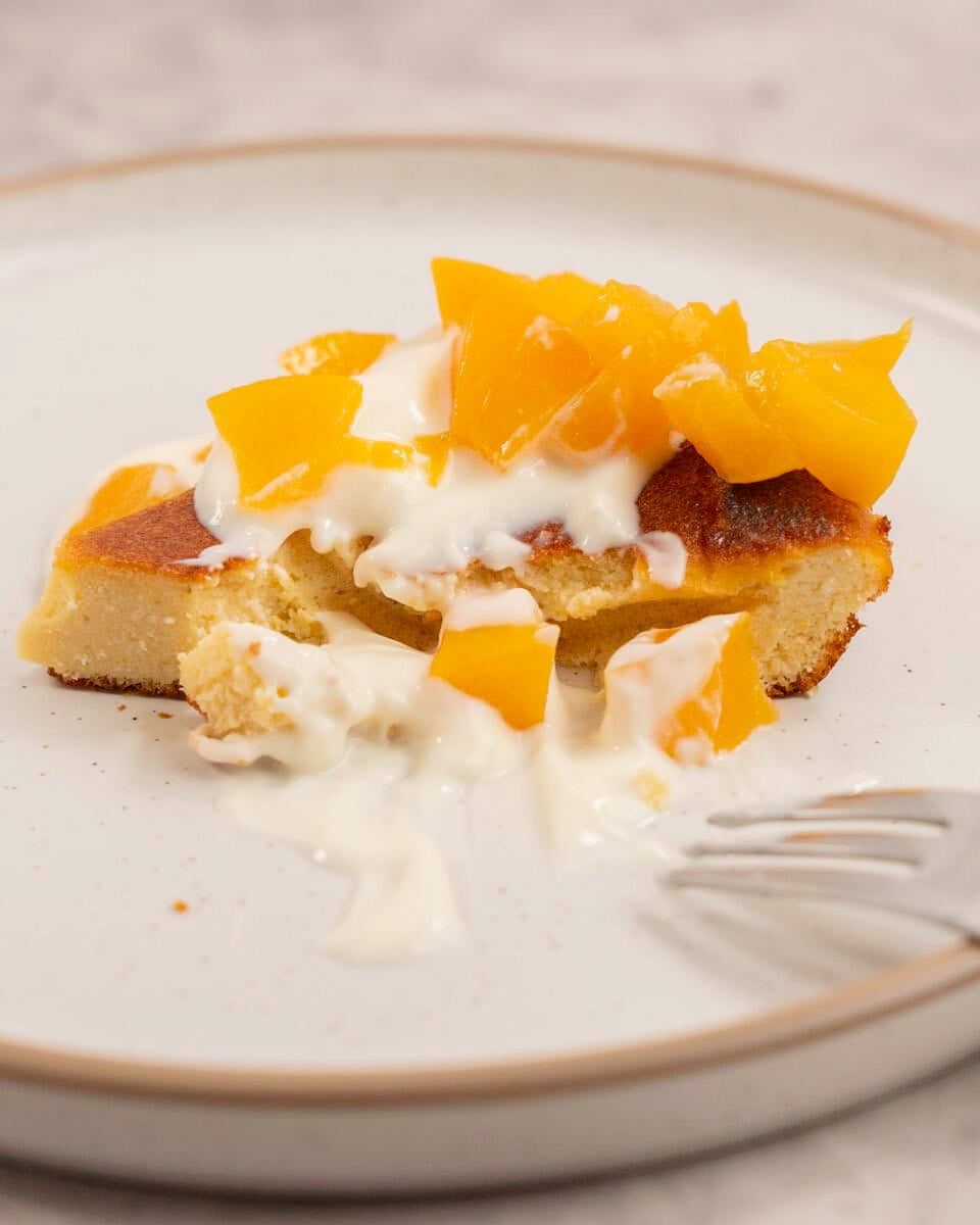 A slice of peach clafoutis on a dessert plate topped with yogurt and diced peaches, with a forkful removed.