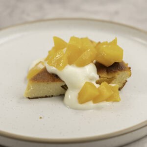 A slice of peach clafoutis on a dessert plate topped with yogurt and diced peaches.