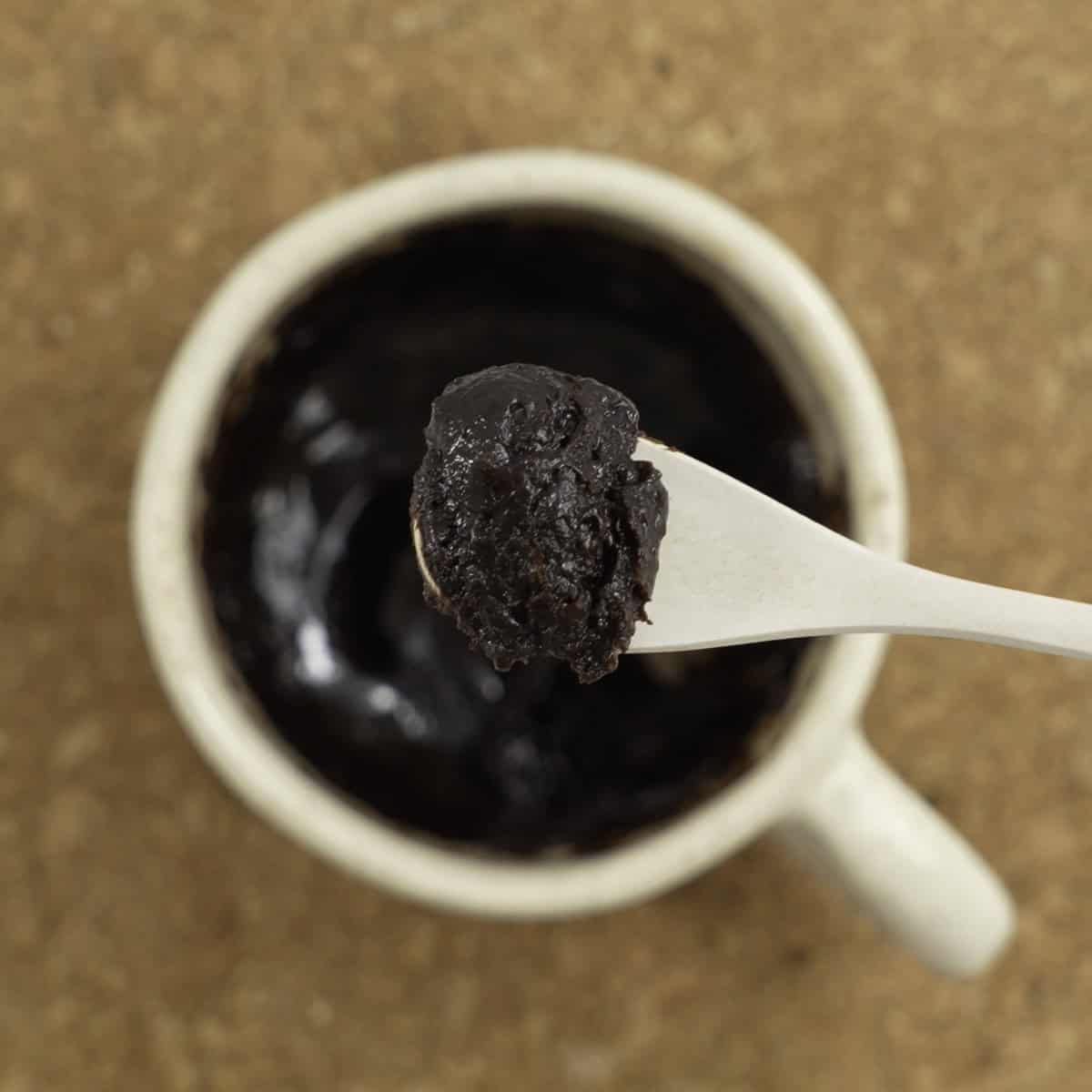 Scoop of brownie on a spoon held up above mug of brownie siting on cork trivet.