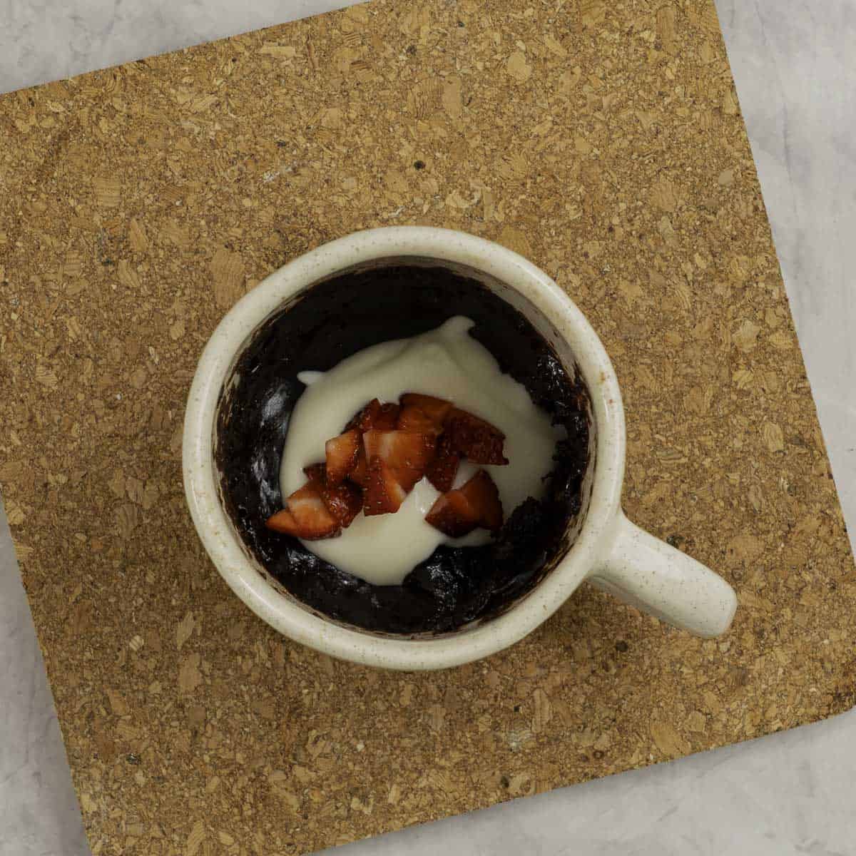Cooked brownie topped with yogurt and chopped strawberries inside a mug sitting on a cork trivet.