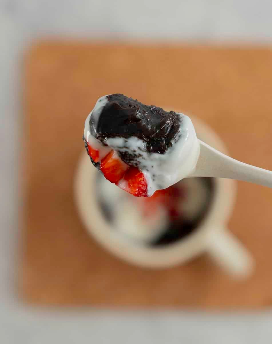 Spoon with brownie, yogurt and strawberries on top held up above mug of brownie.
