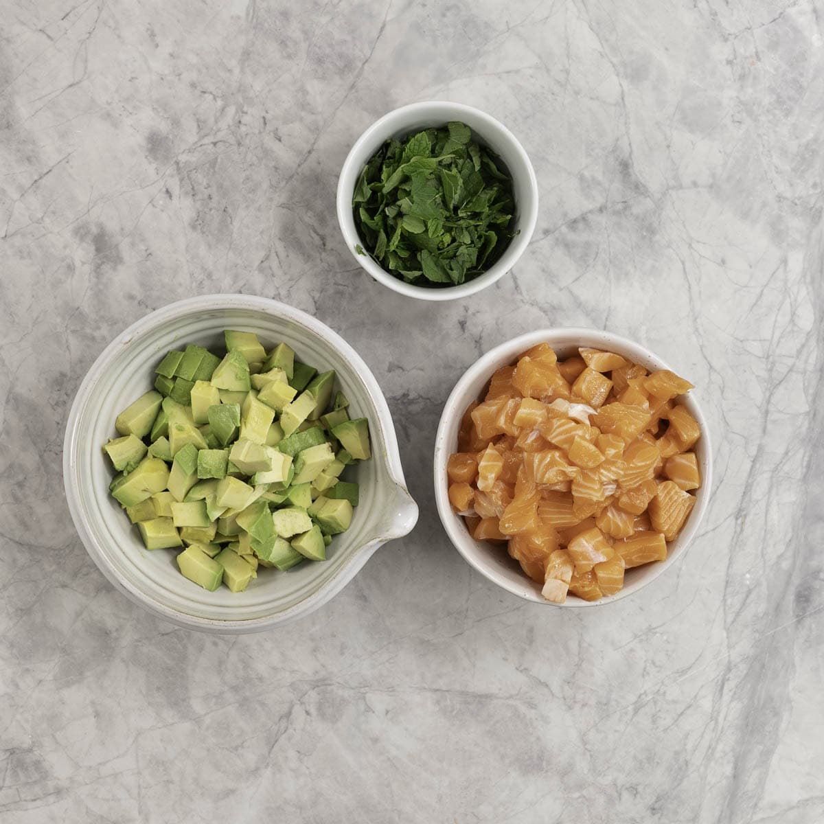 Three bowls containing diced salmon, diced avocado and sliced mint leaves. 