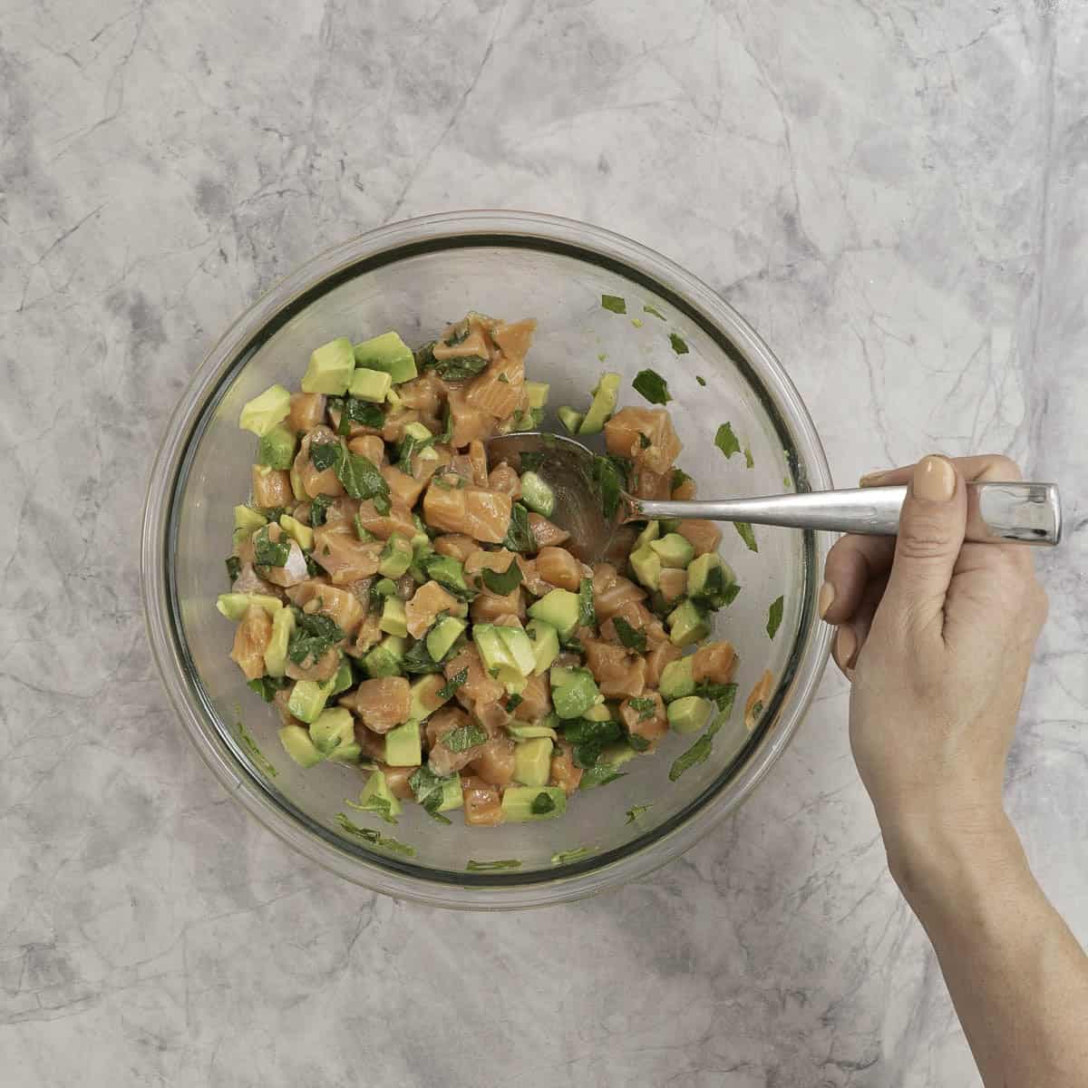 Dressed salmon tartare being spooned from a large glass mixing bowl. 