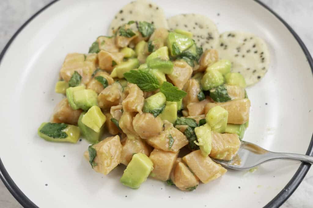 Diced salmon, avocado and mint leaves on a white plate with crisp sesame crackers. 